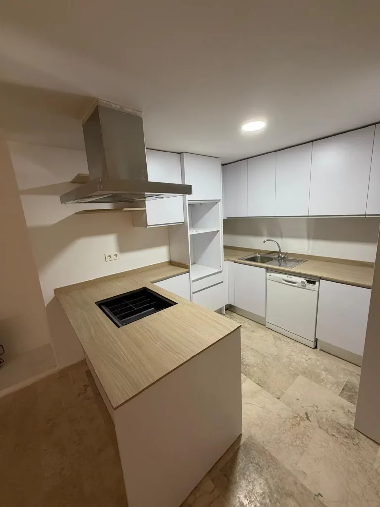 Cocina moderna de diseño con isla central, encimera efecto madera, muebles blancos de acabado liso, campana extractora de acero inoxidable, placa de inducción, fregadero y lavavajillas integrados. Espacio luminoso y funcional, ideal para viviendas contemporáneas.